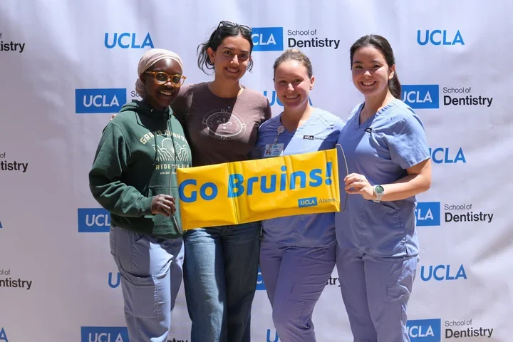 Four smiling dental students, holding a yellow sign reading "Go Bruins!"