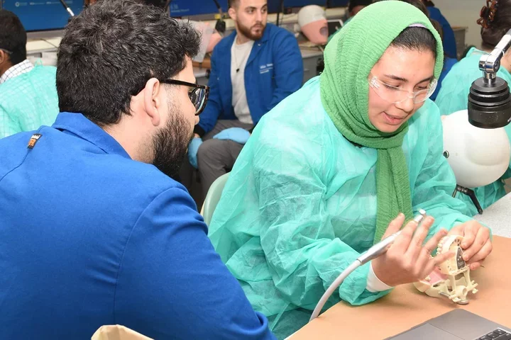 A dental student in blue coat instructing another student in green gown