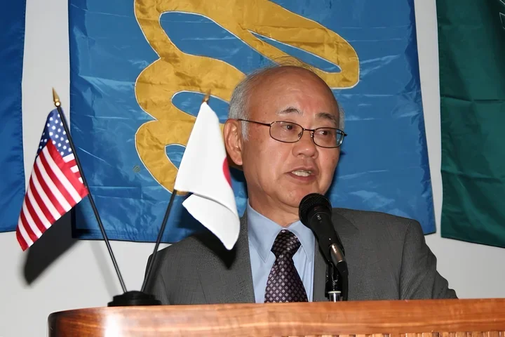 Dr. Henry Takei speaking at a podium, with small USA and Japanese flags next to him and a UCLA banner behind him.