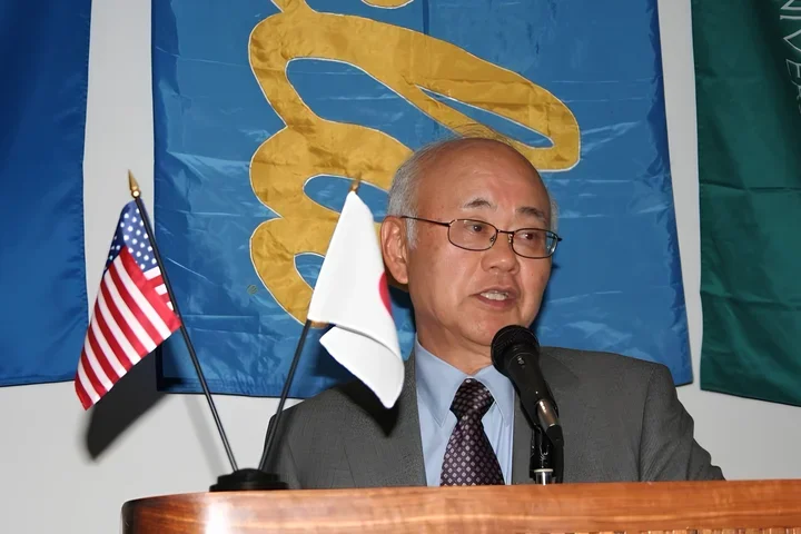 Dr. Henry Takei speaking at a podium, with small USA and Japanese flags next to him and a UCLA banner behind him.