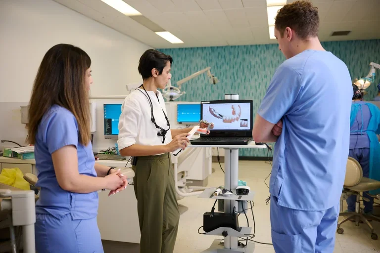 Instructor demonstrates intraoral scanning to two dental students in blue scrubs