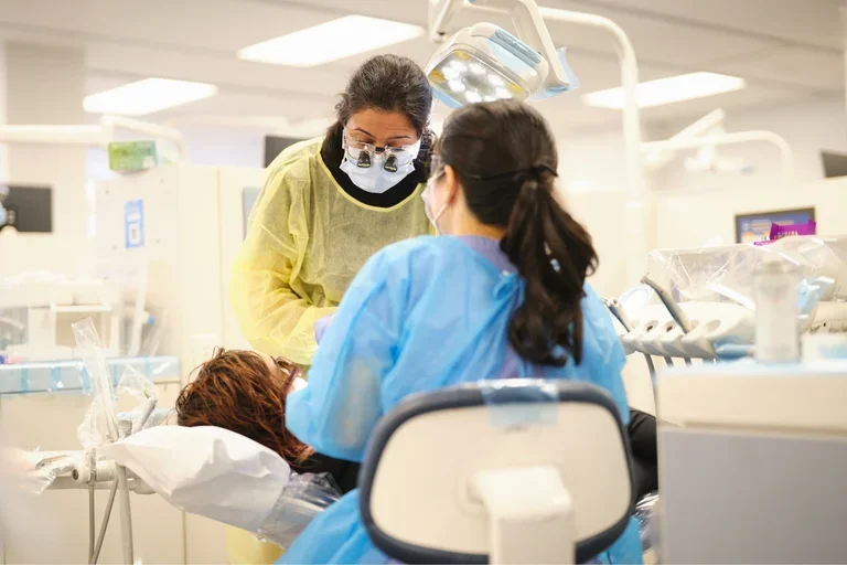 A dentist in yellow gown and dental student in blue gown treating a patient