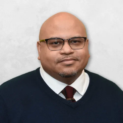 Aaron Shearer is wearing a navy blue sweater over a white shirt with a maroon tie. He is also wearing glasses