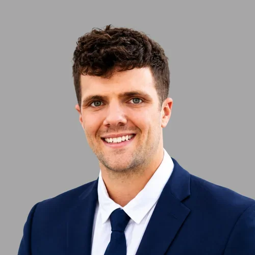 Rance Lockhart smiling, with short brown hair, wearing a navy suit, white shirt, and dark tie against a gray background.