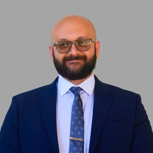 Shahin Arab with a bald head, glasses, and a full beard, wearing a navy suit, white shirt, and patterned tie against a gray background.