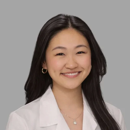 Angelina YueYang Ge smiling, with long dark hair, wearing a white coat and delicate necklace against a gray background.