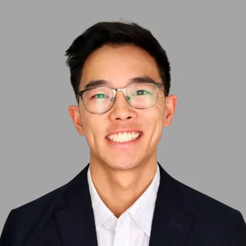 of Lucas Peng smiling, with short dark hair and glasses, wearing a dark suit jacket and white shirt against a gray background.