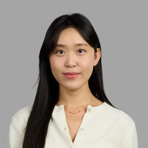 Sun Ha Park with long straight dark hair, wearing a light blouse and necklace against a gray background.