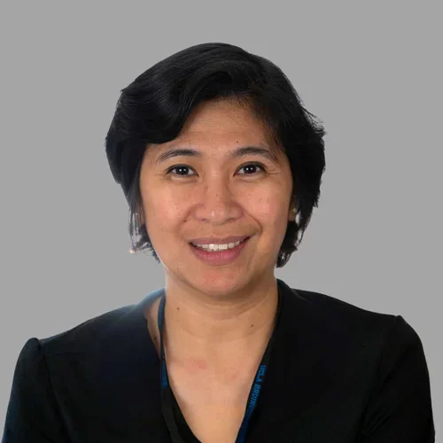 Alma Constantino smiling, with short dark hair, wearing a black blazer against a gray background.