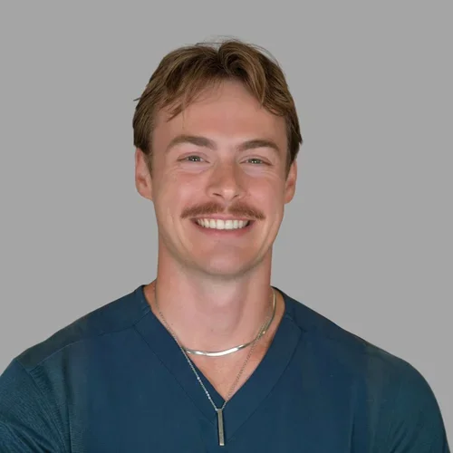 Austin Smith smiling, with light brown hair and a mustache, wearing teal scrubs and a necklace against a gray background.