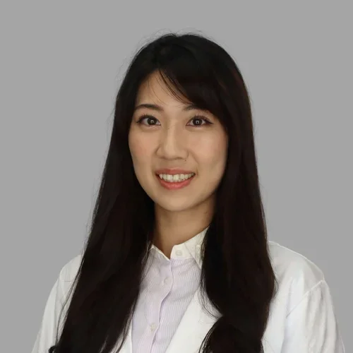 Kristi Lan smiling, with long straight dark hair, wearing a white coat against a gray background.