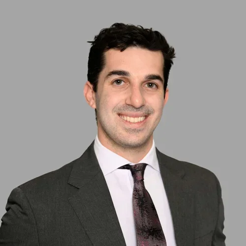 Ryan Neydavood smiling, with short curly dark hair, wearing a gray suit, white shirt, and dark tie against a gray background.