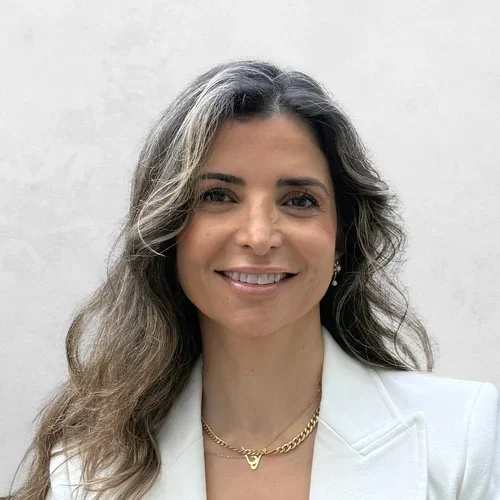 Dr. Rana Al-Rasheed is wearing a white blazer and a gold chain necklace with a small pendant in the shape of a "V" or similar symbol.