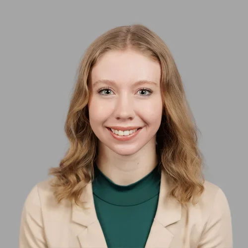 Christina Lindberg smiling, with shoulder-length wavy blonde hair, wearing a beige blazer over a green top against a gray background.
