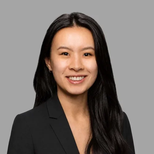 Michelle Chen smiling, with long wavy dark hair, wearing a black blazer against a gray background.