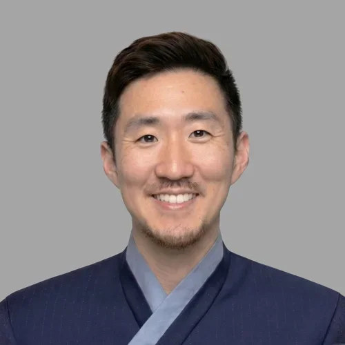 Paul Chung smiling, with short dark hair and light facial hair, wearing a blue coat against a gray background.