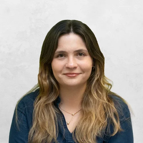 Bruna Fulco wearing a dark blue blouse or shirt. She also has on a delicate gold necklace with two small drop-like pendants and small hoop earrings.