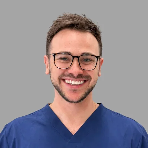 Ariel Oren smiling, with short brown hair, glasses, and a trimmed beard, wearing blue scrubs against a gray background.