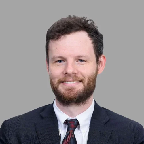 Spencer Austin smiling, with short brown hair and a beard, wearing a dark suit, white shirt, and patterned tie against a gray background.