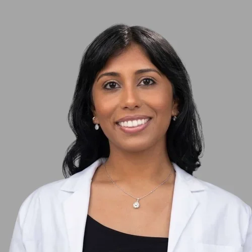 Shalini Sharma smiling, with shoulder-length dark hair, wearing a white coat over a dark top and a necklace against a gray background.