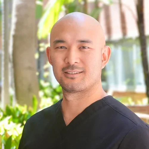 Dr. Grant Matsuura in black dental scrubs, standing in front of a garden
