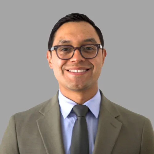 Orlando Ortiz smiling, with short dark hair and glasses, wearing a gray suit, light shirt, and tie against a gray background.