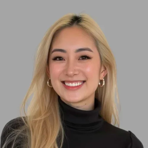 Sammy Gong smiling, with long blonde hair, wearing a black turtleneck and hoop earrings against a gray background.