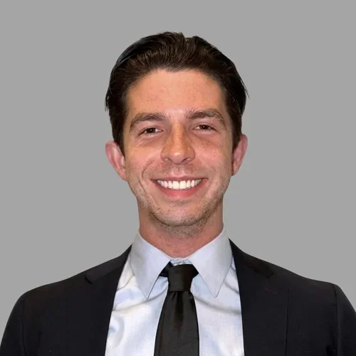 Michael Rehmann smiling, with short brown hair, wearing a dark suit, white shirt, and tie against a gray background.