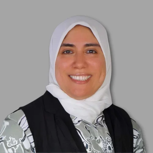 Yomna Nassar smiling, wearing a light-colored hijab and a black vest over a patterned top against a gray background.