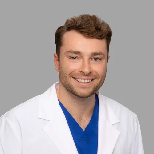 Adam Fuller smiling, with short brown hair and light facial hair, wearing a white coat over blue scrubs against a gray background.
