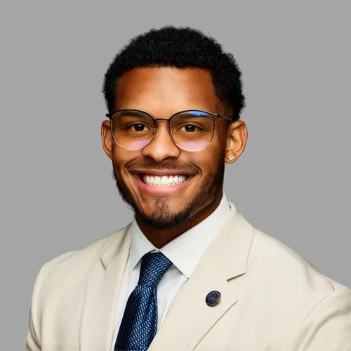 Justin Pratt smiling, with short dark hair, glasses, and a trimmed beard, wearing a light suit jacket, white shirt, and blue tie against a gray background.