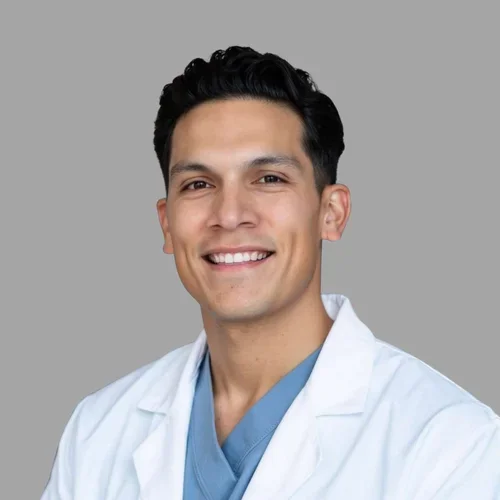 Ali Hernandez smiling, with short dark hair, wearing a white coat over blue scrubs against a gray background.