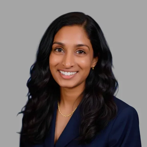 Roslyn Joseph smiling, with long wavy dark hair, wearing a navy blazer and a necklace against a gray background.
