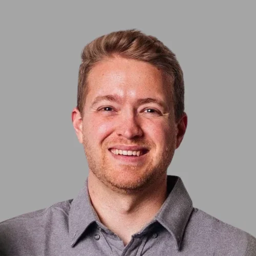 Arick Christopher smiling, with short reddish-brown hair, wearing a gray collared shirt against a gray background.