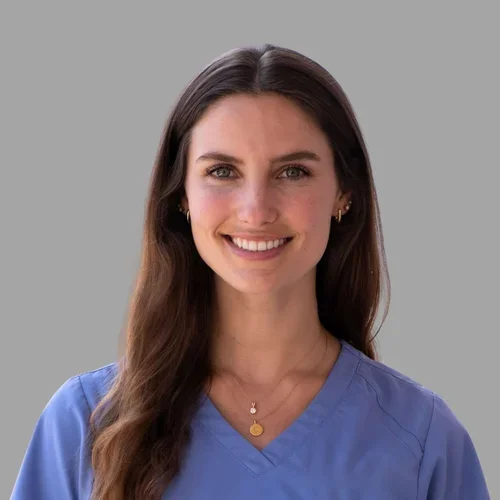 Caitlin Mispagel smiling, with long brown hair, wearing blue scrubs against a gray background.