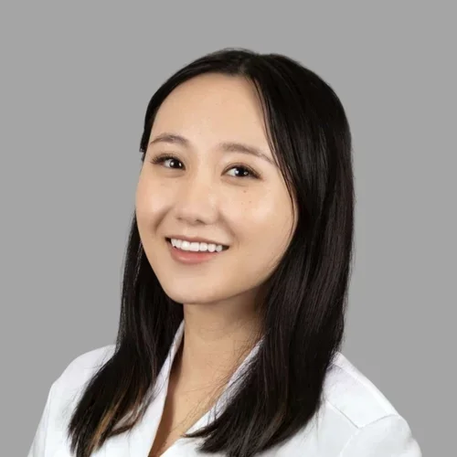 Chelsey Chen smiling, with straight shoulder-length dark hair, wearing a white coat against a gray background.