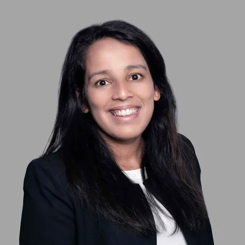 Shveta Duggal smiling, with long dark hair, wearing a dark blazer over a light top against a gray background.