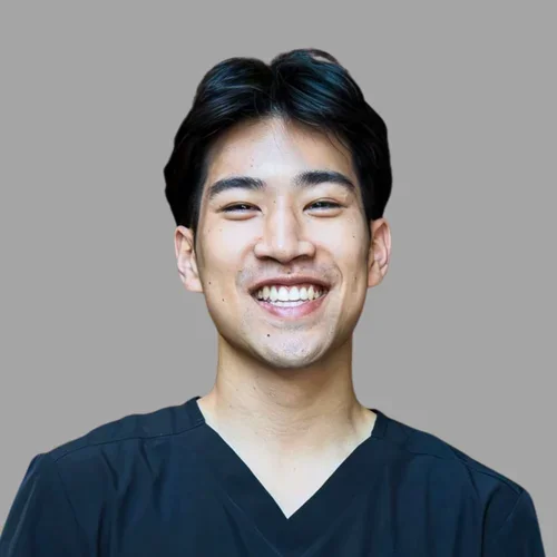 Brendan Co smiling, with short dark hair, wearing dark scrubs against a gray background.