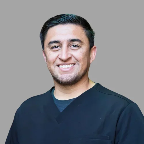 Christopher Orellana smiling, with short dark hair, wearing dark scrubs against a gray background.