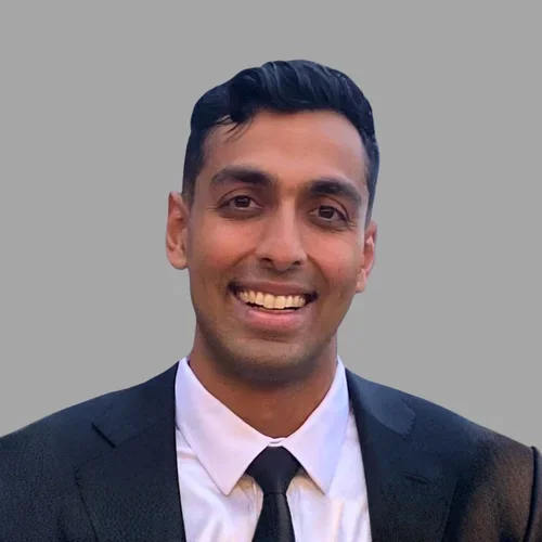 Rohan Mahadevia smiling, with short dark hair, wearing a dark suit, light shirt, and tie against a gray background.