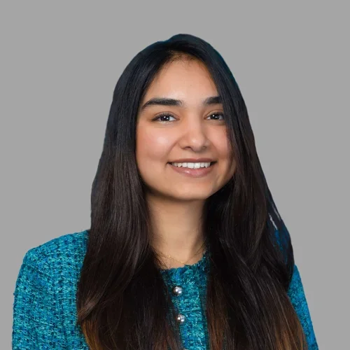 of Mishri Parikh smiling, with long dark hair, wearing a teal textured top against a gray background.