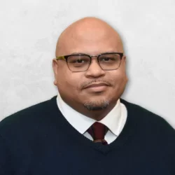 Aaron Shearer is wearing a navy blue sweater over a white shirt with a maroon tie. He is also wearing glasses