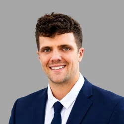 Rance Lockhart smiling, with short brown hair, wearing a navy suit, white shirt, and dark tie against a gray background.