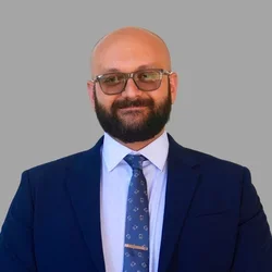 Shahin Arab with a bald head, glasses, and a full beard, wearing a navy suit, white shirt, and patterned tie against a gray background.