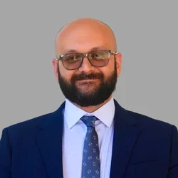Shahin Arab with a bald head, glasses, and a full beard, wearing a navy suit, white shirt, and patterned tie against a gray background.