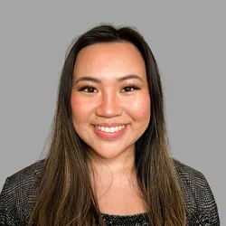 Caroline Hoang smiling, with long brown hair, wearing a dark textured jacket over a black top against a gray background.