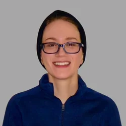 Medina Kerdjani, smiling, wearing glasses, a black headscarf, and blue jacket against a gray background.