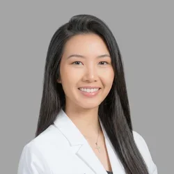 Gia Nguyen smiling, with long straight dark hair, wearing a white coat against a gray background.