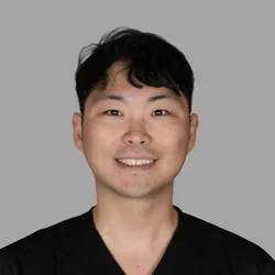 of Ray Kim smiling, with short dark hair, wearing black scrubs against a gray background.