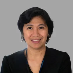 Alma Constantino smiling, with short dark hair, wearing a black blazer against a gray background.